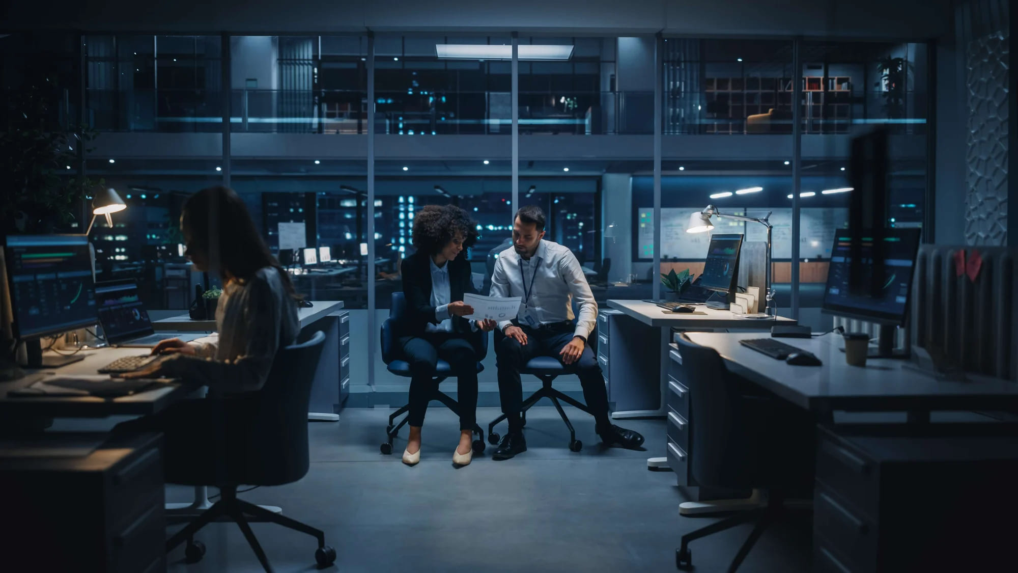 Two people sitting at desks in an office at night.